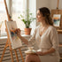 A woman in a light dress smiles, painting a colorful abstract piece while wearing a delicate gold-filled y-necklace in a bright, cozy room.